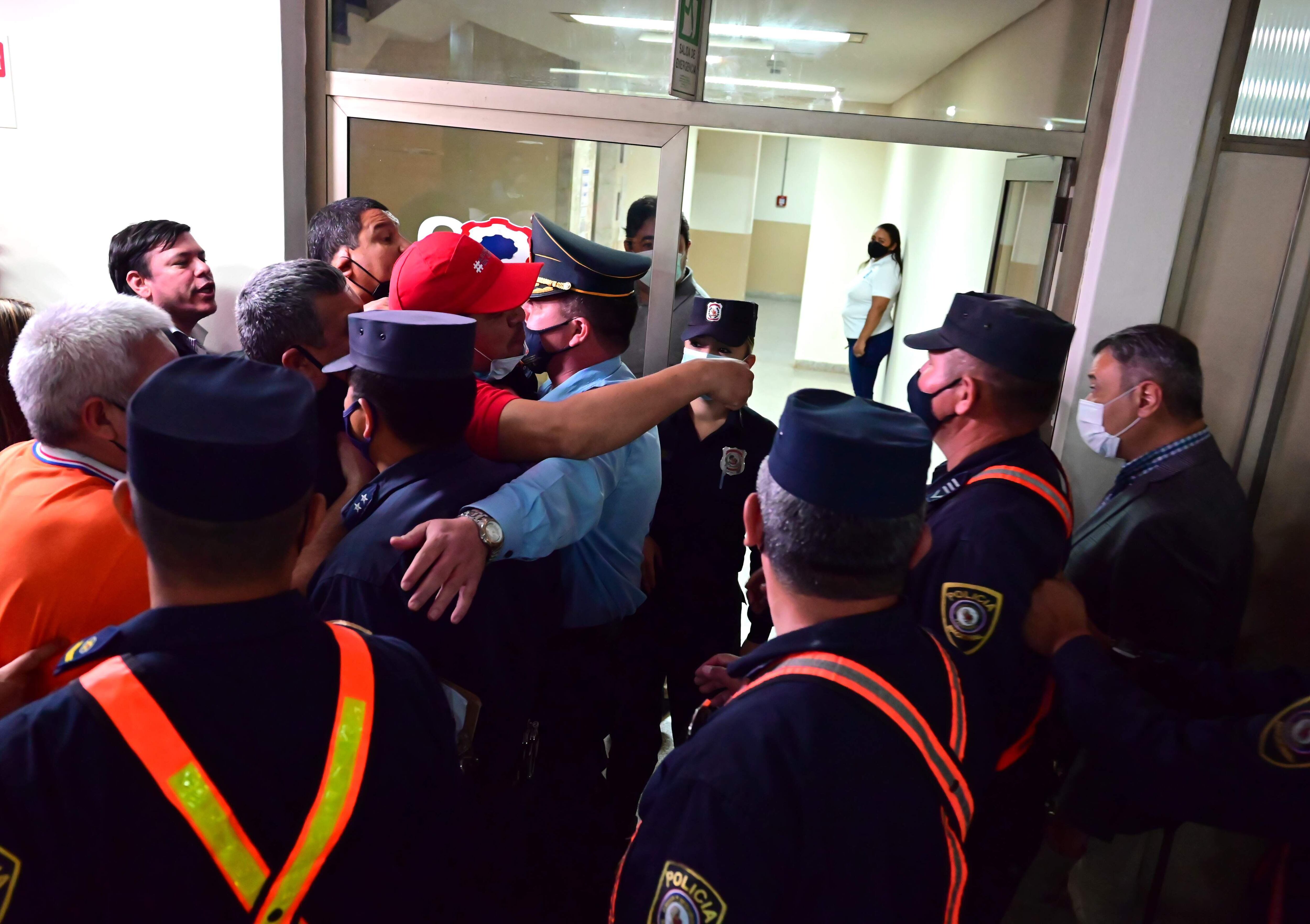 A la derecha, custodiado por policías se retira Humberto Blasco, presidente de la Junta asuncena. A la izquierda los funcionarios. Uno de ellos le grita y señala (quepi rojo) y posteriormente rompe el vidrio de la puerta. 