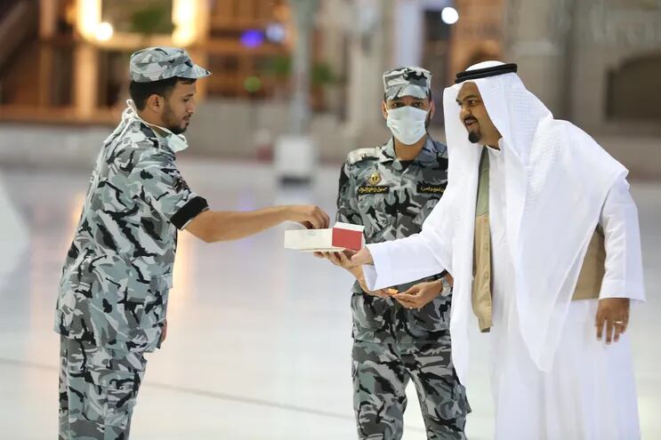 Un fiel musulmán saudí ofrece golosinas a guardias de seguridad, uno de ellos con mascarilla, en la Gran Mezquita de La Meca.