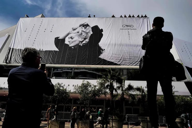 Transeúntes observan cómo los obreros despliegan el cartel oficial de la 76° edición del Festival de Cannes, que tiene como protagonista a la actriz Catherine Deneuve en una fotografía de Jack Garofalo. El Palacio del festival recibirá desde mañana a varias estrellas del séptimo arte.