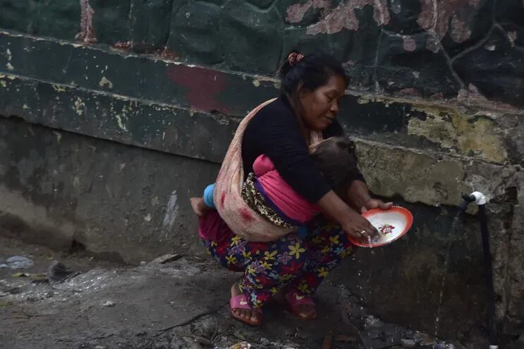 Mujer indígena lava platos entre agua servida en la acera de la Intendencia Militar donde funciona el Indi, sobre la avenida Artigas, mientras carga a su hija.