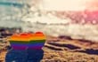 Dos corazones con los colores del arcoíris incrustados en la arena de una playa, frente a un curso de agua.