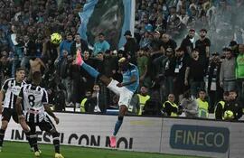 Víctor Osimhen puntea el balón ante dos jugadores rivales y con una imagen de Diego Maradona en el fondo, en las tribunas del estadio de Udinese, durante el partido de ayer que coronó campeón al Napoli.