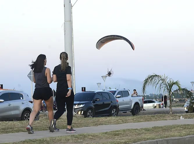 Un parapente hace un vuelo bajo, mientras dos jovenes realizan su actividad de caminata en la Costanera.
