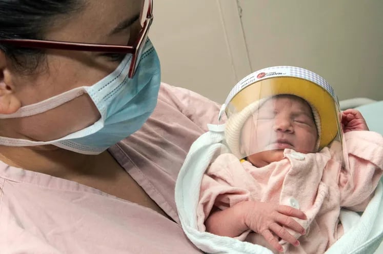 Una mujer con mascarilla sostiene a su bebé recién nacido, que usa un protector facial.