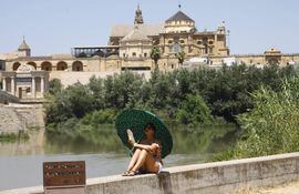 Una turista se protege del calor con un paraguas y un abanico a la orilla del río Guadalquivir cerca de la Mezquita-Catedral de Córdoba ( al fondo).