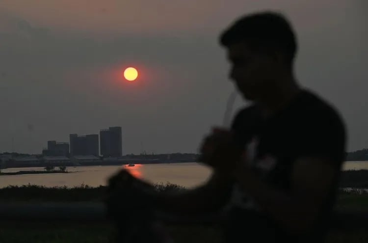 Un hombre aplaca el intenso calor tomando tereré en la Costanera de Asunción. De fondo se ve la silueta de los nuevos edificios del gobierno en la Bahía de Asunción, durante una puesta de sol.