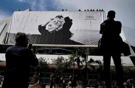 Transeúntes observan cómo los obreros despliegan el cartel oficial de la 76° edición del Festival de Cannes, que tiene como protagonista a la actriz Catherine Deneuve en una fotografía de Jack Garofalo. El Palacio del festival recibirá desde mañana a varias estrellas del séptimo arte.