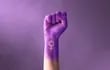Un puño en alto de un brazo de mujer, teñido de lila, con el símbolo del feminismo.