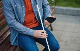 Una persona sentada en un banco público, que tiene un bastón blanco en una de sus manos y en la otra sostiene un teléfono celular.