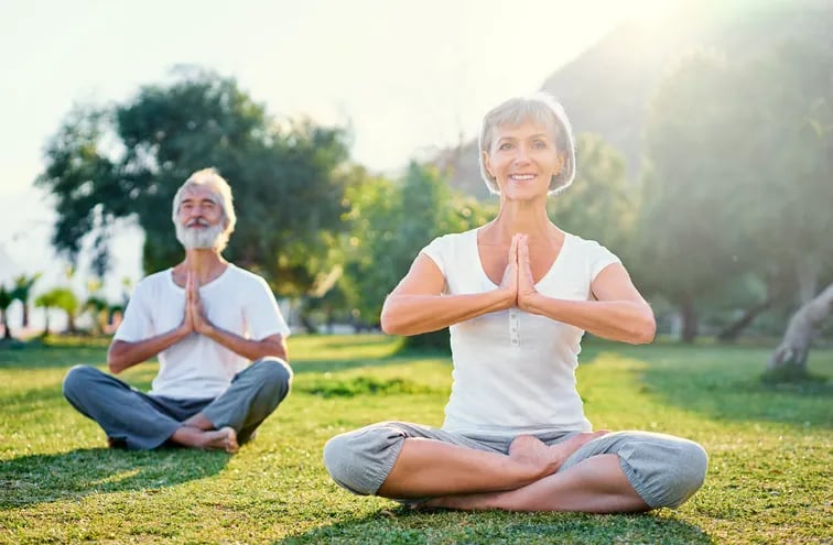 Una pareja de adultos mayores practica yoga al aire libre, sobre el pasto, con rostros complacidos.
