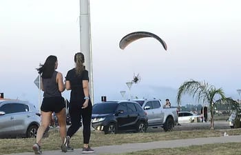 Un parapente hace un vuelo bajo, mientras dos jovenes realizan su actividad de caminata en la Costanera.