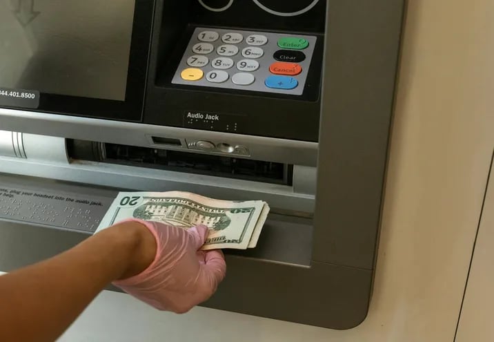Se observa el brazo de una persona y en su mano tiene unos dólares al frente de unos cajeros automáticos.