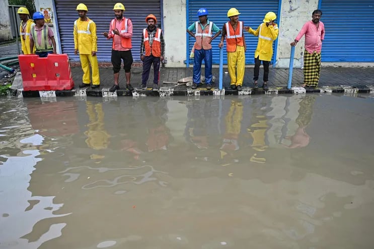 Hombres de pie junto a una acera a lo largo de una calle inundada después de las lluvias, mientras se espera que el ciclón Michaung toque tierra.