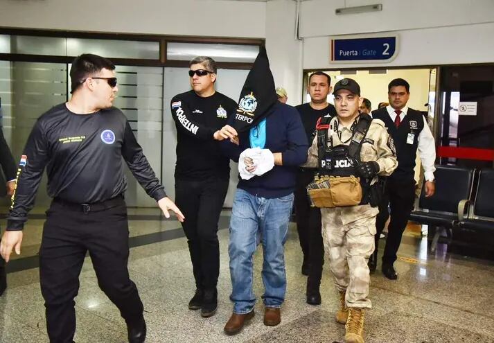 Miguel Ángel Insfrán (con la cabeza cubierta) bajo custodia de agentes de Interpol y la Policía Nacional al momento de su llegada a Paraguay, este viernes en el aeropuerto internacional Silvio Pettirossi.