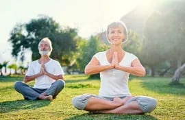 Una pareja de adultos mayores practica yoga al aire libre, sobre el pasto, con rostros complacidos.