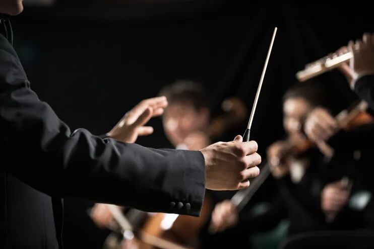 Brazos de un director de orquesta. Con la mano derecha sostiene la batuta. En el fondo difuminado hay músicos tocando el violín, flauta y otros instrumentos.