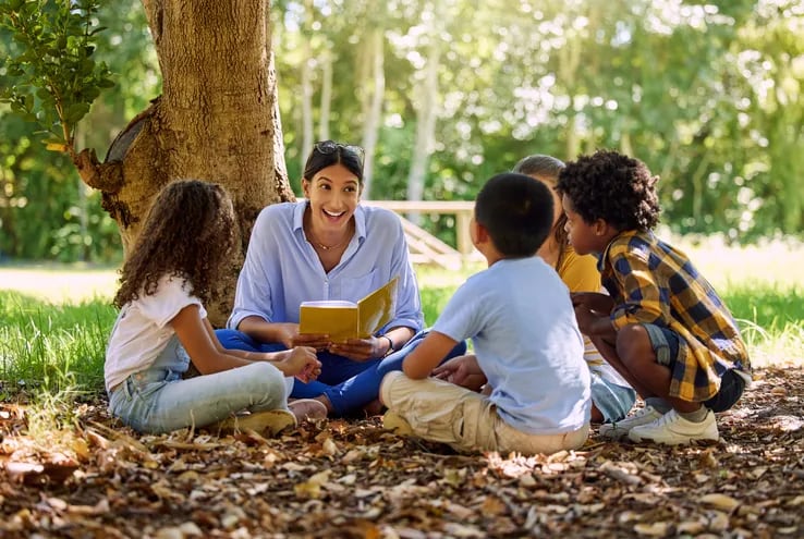 Una mujer adulta sonríe y lee un cuento a dos niñas y dos niñas sentados a su alrededor, sobre hojas secas, a la sombra de un árbol.