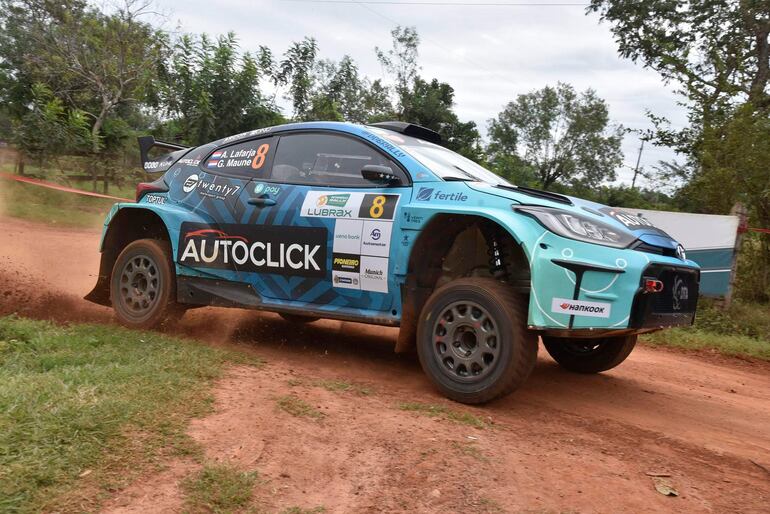 Coche de rally azul y negro número 8 en movimiento, levantando polvo en un camino de tierra rodeado de vegetación.