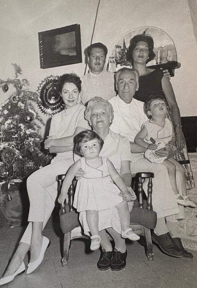 Mujer mayor sentada con tres niñas, todos sonrientes; ambiente decorado con un árbol de Navidad y otros elementos festivos.