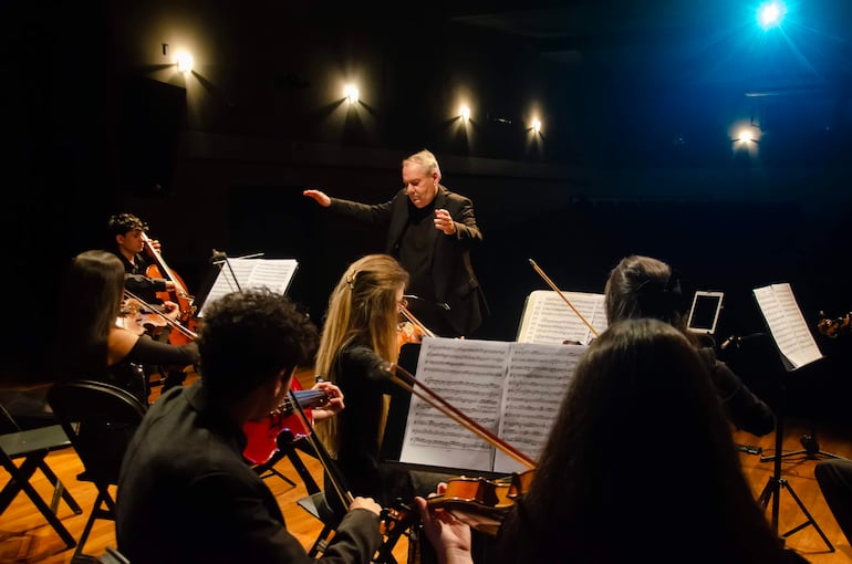 Miguel Ángel Gilardi dirige a la Orquesta de Cámara Juvenil, con músicos tocando violonchelo y violines en un ambiente iluminado.