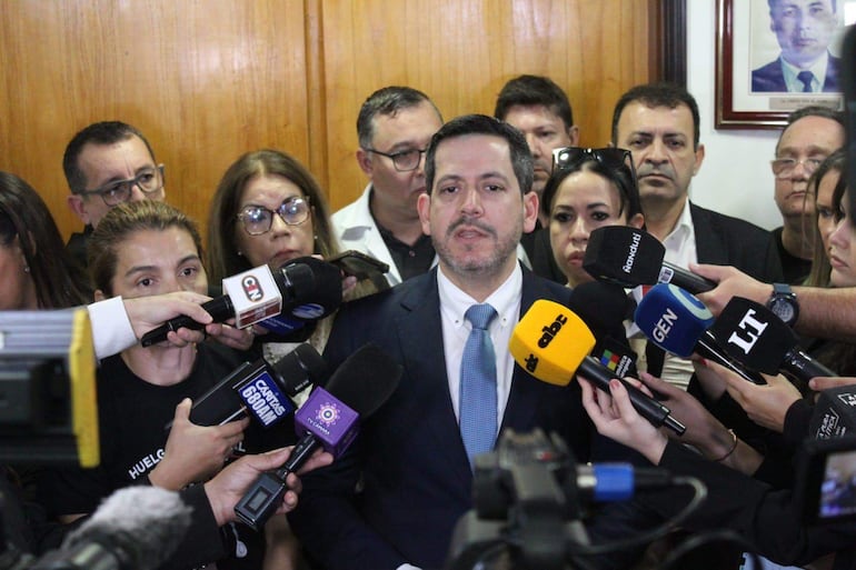 Raul Luis Latorre Martínez en traje oscuro y corbata clara, hablando a periodistas, rodeado de asistentes atentos con micrófonos.