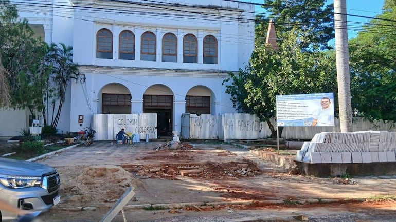La imagen presenta un entorno exterior con un edificio blanco que parece ser de utilidad pública, con varias ventanas enmarcadas en madera. . 

La escena muestra un espacio en construcción con tierra y bloques apilados en el suelo, lo que indica actividad de obras.