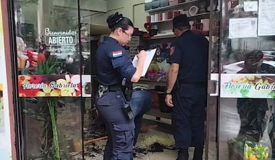 Mujer policía anotando en libreta, mientras otro policía observa atento en la florería Gabriela, rodeados de flores y decoración del local.
