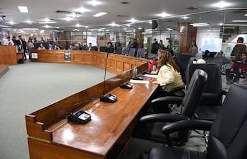 Mujer sentada en una mesa con micrófono, rodeada de una audiencia atenta en sala con decoración de madera y moqueta verde.