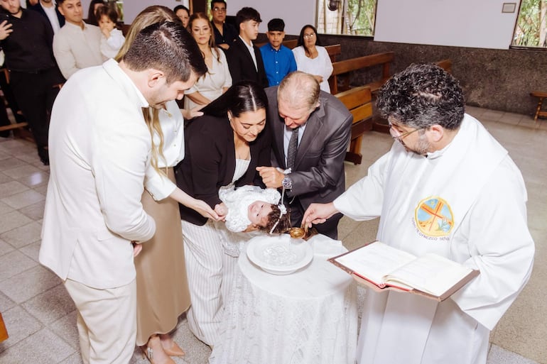 Sacerdote en vestimenta litúrgica sostiene un libro y agua, mientras una mujer viste al bebé en un vestido blanco durante la ceremonia.