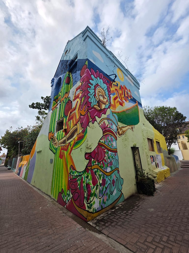 Mural colorido en edificio de esquina, sin personas, mostrando figuras humanas estilizadas y un entorno urbano luminoso.