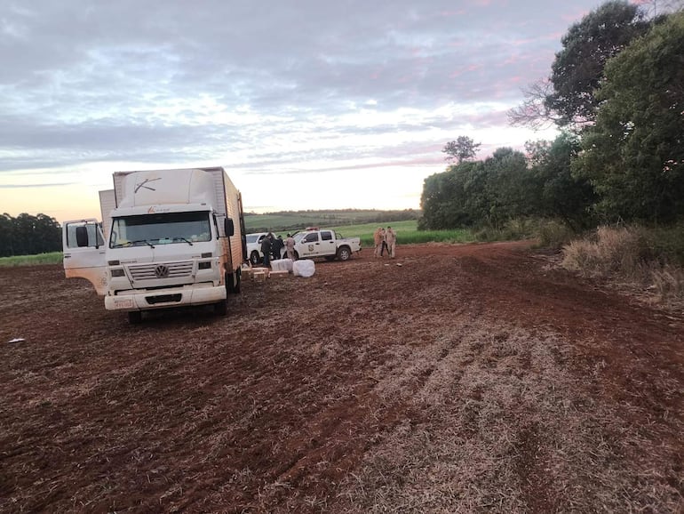 Cuatro personas, entre ellas funcionarios, revisan cajas en el suelo cerca de un camión y una camioneta en un campo abierto.