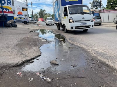 Charco de agua sucia reflejando el cielo, con vehículos en movimiento y desechos visibles en el suelo.