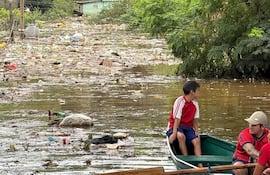 Niño con camiseta roja y pantalones cortos azules en canoa, junto a hombre con gorra, navegando en agua llena de basura.