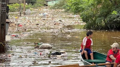 Niño con camiseta roja y pantalones cortos azules en canoa, junto a hombre con gorra, navegando en agua llena de basura.