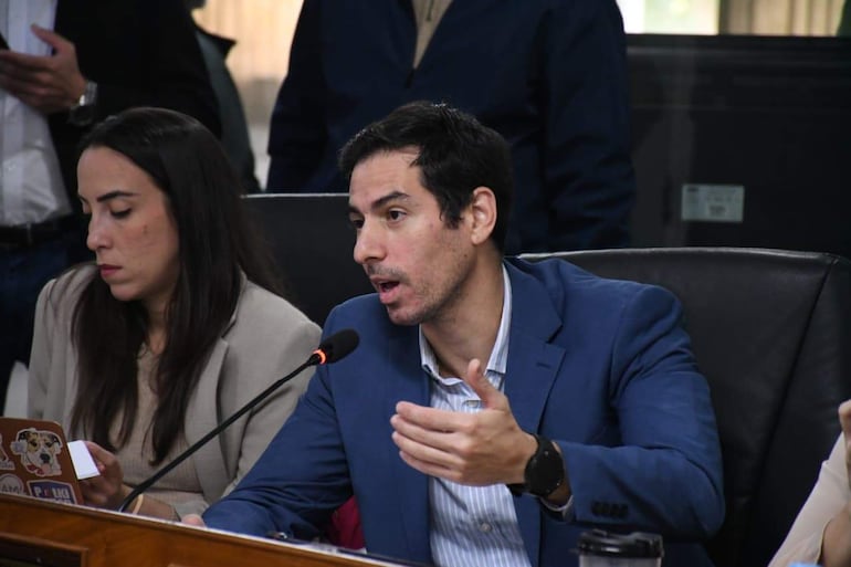 Álvaro Grau en traje azul gesticulando, mientras Jazmín Galeano toma notas, en ambiente de sala de reuniones.
