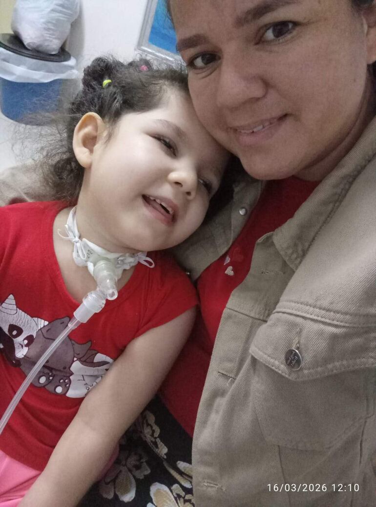La pequeña con tubo en el cuello sonríe apoyada en su madre, a pesar de problema que posee.