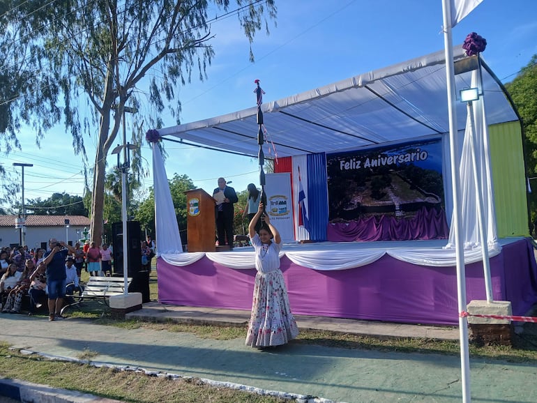 Mujer en vestido de gala muestra un estandarte, mientras un hombre vestido con saco habla en el escenario.