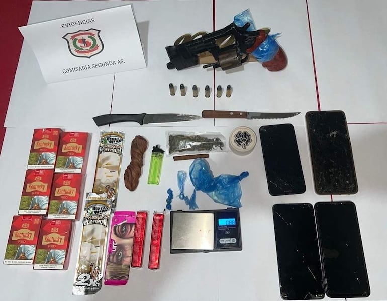 Objetos incautados sobre mesa blanca: revólver, cuchillos, balanza y teléfonos móviles en ambiente policial.