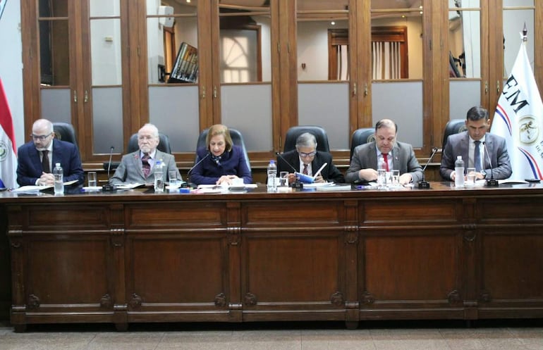 César Garay Zuccolillo en reunión formal, acompañado por Alicia Pucheta y otros, todos en atuendos elegantes y en actitud concentrada.