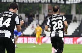 Jugadores Alexis Cecon y Fernández en acción, vistiendo camisetas blancas y negras en un campo de fútbol con ambiente festivo.