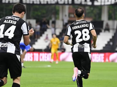 Jugadores Alexis Cecon y Fernández en acción, vistiendo camisetas blancas y negras en un campo de fútbol con ambiente festivo.