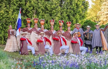 Catorce personas lucen trajes tradicionales paraguayos, sonriendo y sosteniendo instrumentos musicales, en un entorno natural.