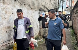 Dos hombres, uno con chaqueta gris y bolsa de tomates, otro con camiseta azul y ramo de papas, caminan en un pasillo urbano apretado.
