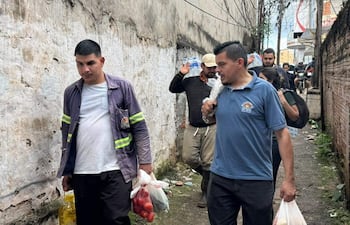 Dos hombres, uno con chaqueta gris y bolsa de tomates, otro con camiseta azul y ramo de papas, caminan en un pasillo urbano apretado.