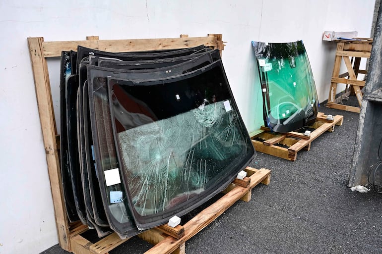 Cristales de automóviles apilados sobre plataformas de madera, algunos con arañazos y grietas, en un taller exterior industrial.