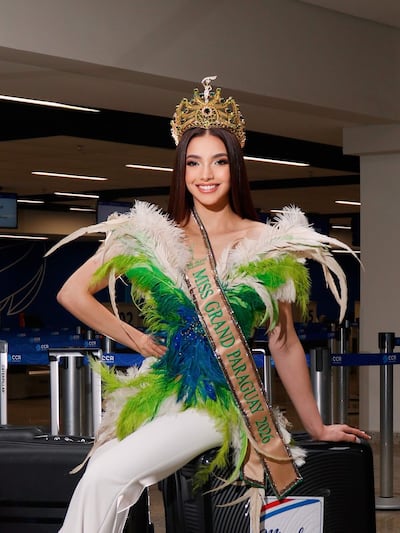 Mujer sonriente con vestido verde y blanco y corona de Miss Grand Paraguay en el aeropuerto, posando sobre una maleta.
