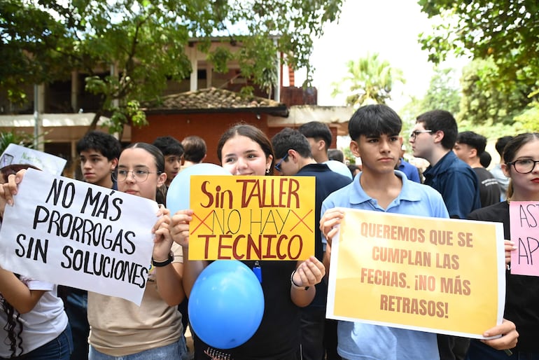 Grupo de jóvenes, algunos con gafas y sosteniendo carteles, expresan demandas frente a un edificio escolar con áreas verdes al fondo.