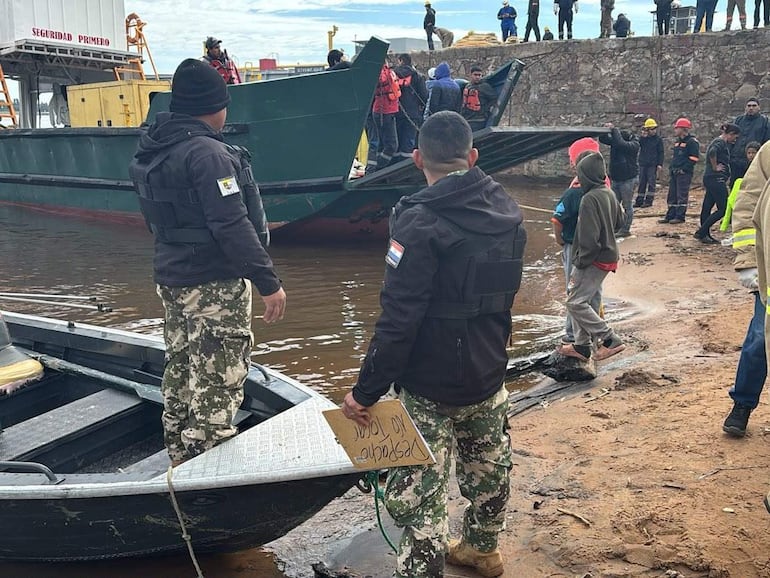 Dos hombres, uno en camuflado y chaleco de seguridad, sostienen un cartel que dice 'Perdido' en un ambiente acuático con cielo nublado.