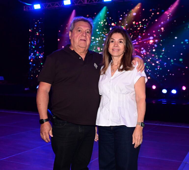Hombre canoso en camiseta negra y mujer en blusa blanca sonríen juntos en ambiente festivo, con luces brillantes al fondo.