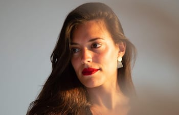 Mujer de cabello largo y suelto, con labios rojos y pendientes geométricos, posando con expresión serena frente a un fondo suave.